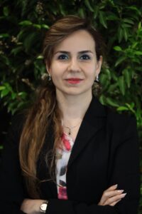 Picture of woman with folded arms, wearing a black jacket over a flowery blouse. She is standing in front of a green bush.