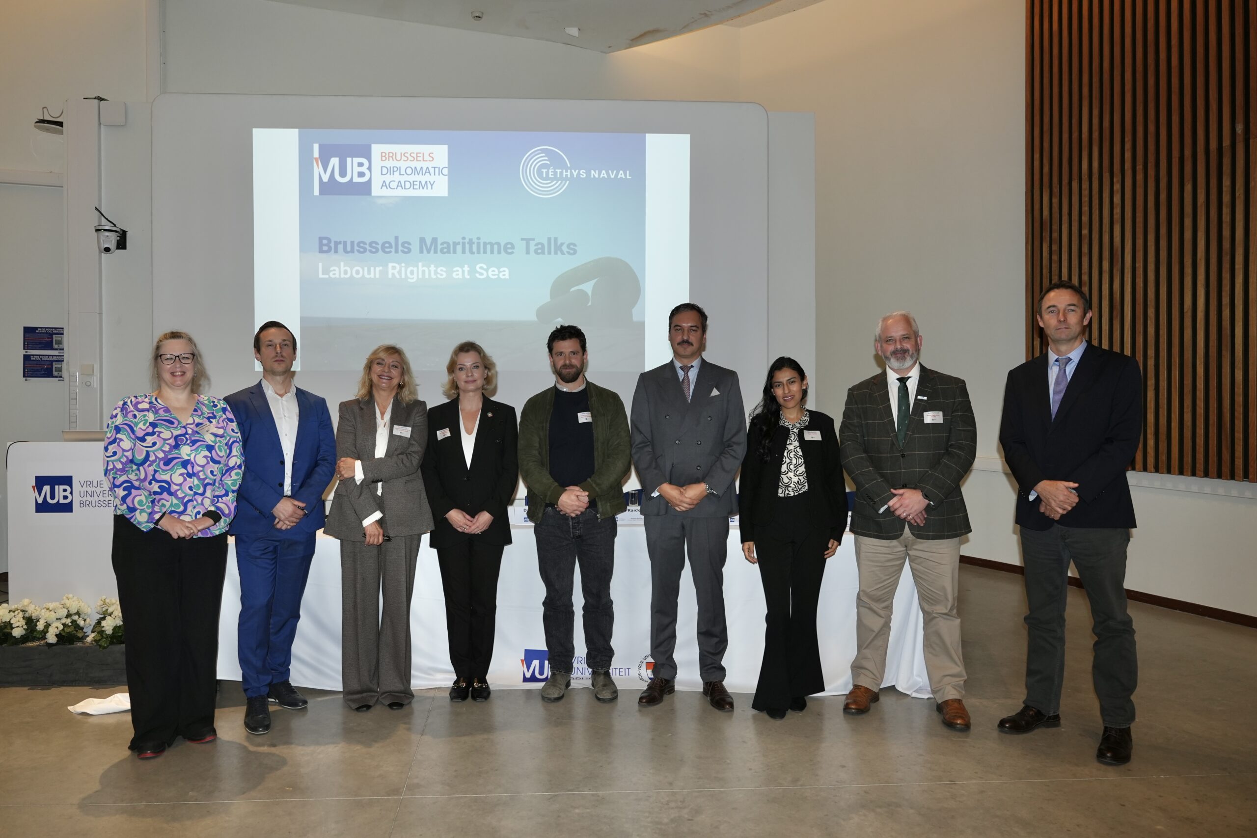 Photograph of 4 women and 5 men standing in front of a screen with logos of the Brussels Diplomatic Academy and Thetys Naval and the words Brussels Maritime Talks Labour Rights at Sea.