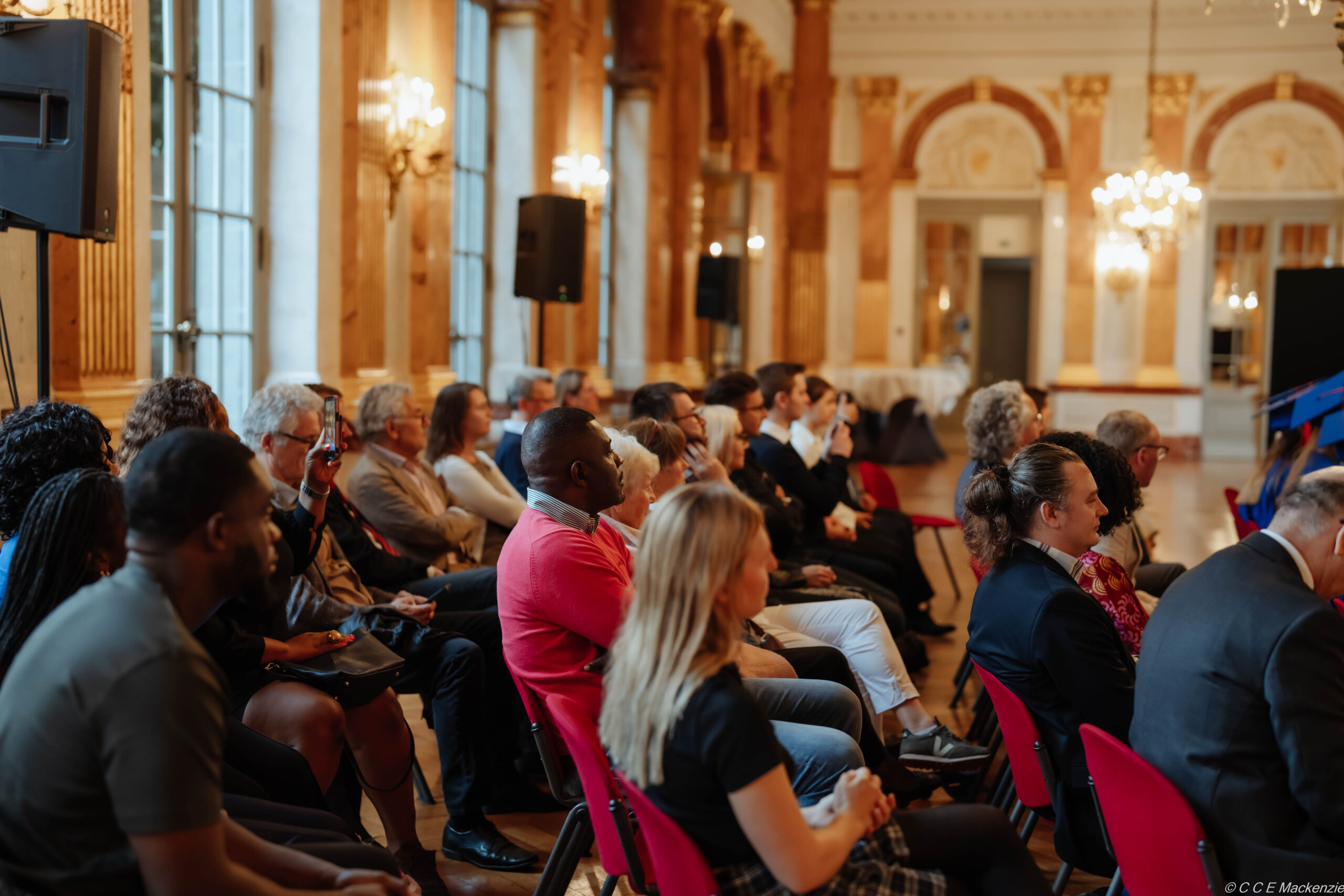 Diverse group of people sitting listening to someone. The room is elegant with high ceiling, tall windows and chandeliers.