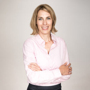 Woman with fair hair, smiling, facing camera, wearing a pink shirt and standing with arms gently crossed
