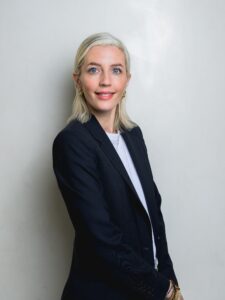 Waist up shot of woman standing side on, head facing the camera, smiling.